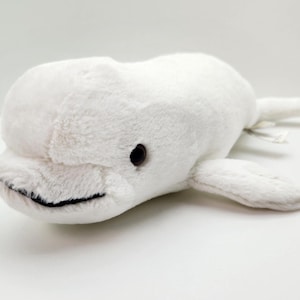 May include: A plush beluga whale toy made of soft white fabric. The stuffed animal features a rounded head, a small black stitched mouth, and two dark brown plastic eyes positioned on the sides of its head, with side fins and a tail.