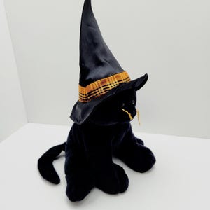 Puede incluir: Un gato de peluche negro con un sombrero de bruja negro con una banda a cuadros dorada y naranja. El gato tiene bigotes amarillos y est&aacute; sentado sobre una superficie blanca. El sombrero es alto y puntiagudo.