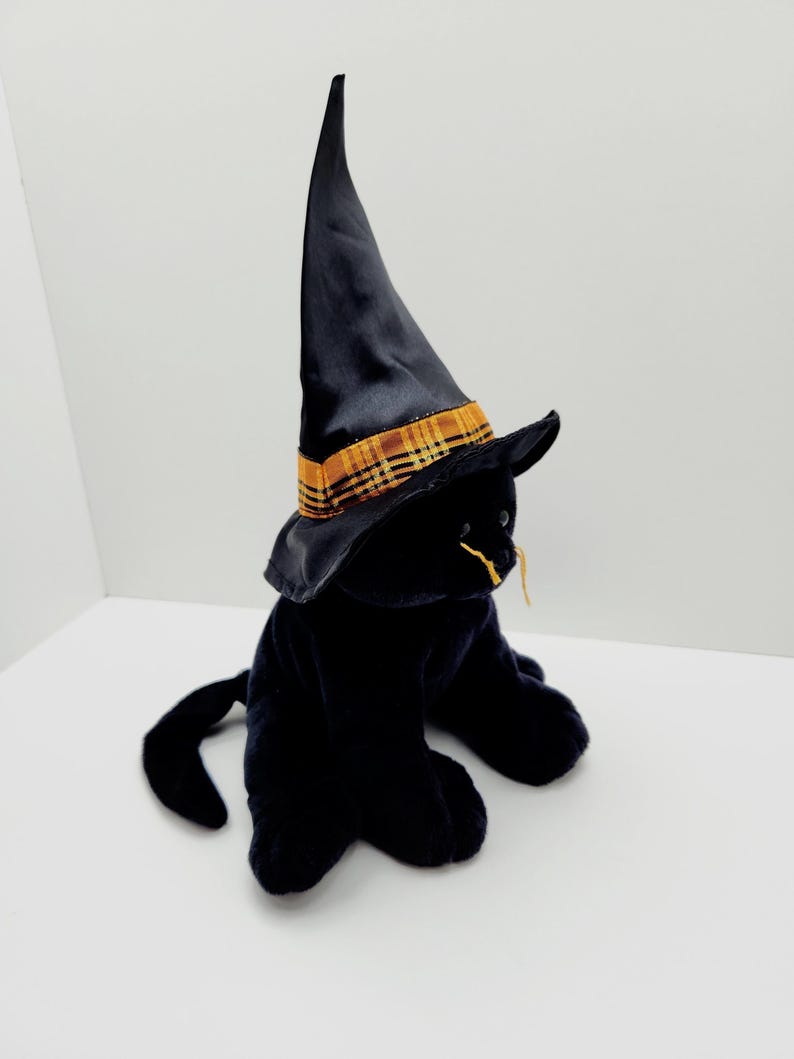 Puede incluir: Un gato de peluche negro con un sombrero de bruja negro con una banda a cuadros dorada y naranja. El gato tiene bigotes amarillos y est&aacute; sentado sobre una superficie blanca. El sombrero es alto y puntiagudo.