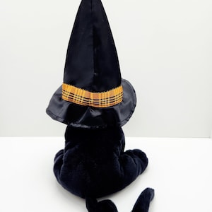Puede incluir: Un gato de peluche negro con un sombrero de bruja negro con una banda a cuadros amarilla y naranja. El gato est&aacute; sentado de espaldas al espectador, con la cola enroscada. El sombrero es alto y puntiagudo, t&iacute;pico de Halloween.