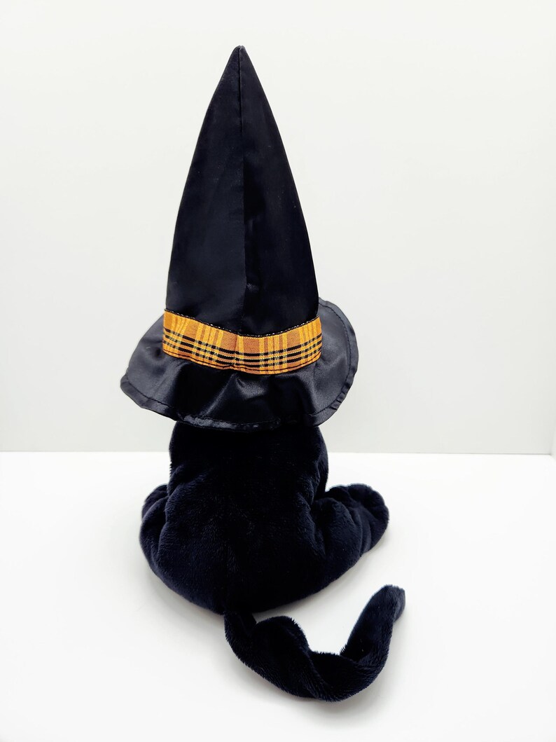 Puede incluir: Un gato de peluche negro con un sombrero de bruja negro con una banda a cuadros amarilla y naranja. El gato est&aacute; sentado de espaldas al espectador, con la cola enroscada. El sombrero es alto y puntiagudo, t&iacute;pico de Halloween.