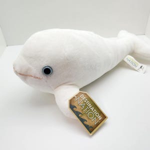 May include: A white plush beluga whale toy with dark blue plastic eyes. A tag attached to the fin reads DESTINATION NATION with a graphic of blue waves. A smaller white fabric tag on the side also displays the text NATION.