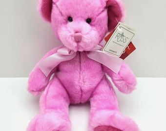 May include: A bright pink plush teddy bear with soft fur, featuring a light pink satin ribbon tied around its neck. The bear has black plastic eyes, a pink stitched nose, and the word LIBRA embroidered in blue on its left foot pad. A white Russ brand tag is attached to the ear.