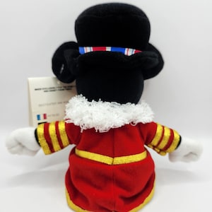 Op de afbeelding: Een pluche Mickey Mouse-speelgoed verkleed als Beefeater, van achteren gezien. Het speelgoed heeft een zwarte hoed met een rood-wit-blauw lint, een rode jas met gouden randen en witte handschoenen. De achtergrond is wit.