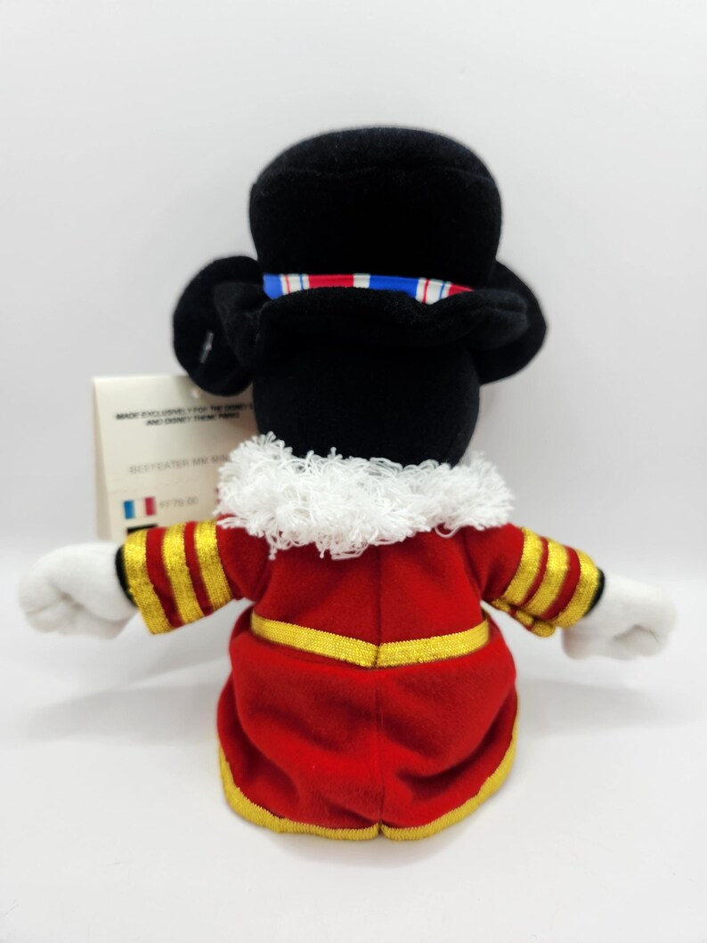 Op de afbeelding: Een pluche Mickey Mouse-speelgoed verkleed als Beefeater, van achteren gezien. Het speelgoed heeft een zwarte hoed met een rood-wit-blauw lint, een rode jas met gouden randen en witte handschoenen. De achtergrond is wit.