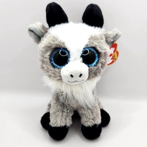 May include: A soft toy goat with a white face and a grey and white body. It has large blue glittery eyes, black horns and hooves, and a white beard. A red tag is attached to the ear.