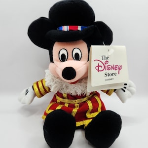 Op de afbeelding: Mickey Mouse pluche speelgoed met een rood en goud kostuum, een witte gefranjerde kraag en een zwarte hoed. Het speelgoed heeft zwarte oren en voeten, witte handschoenen en een Disney Store label.