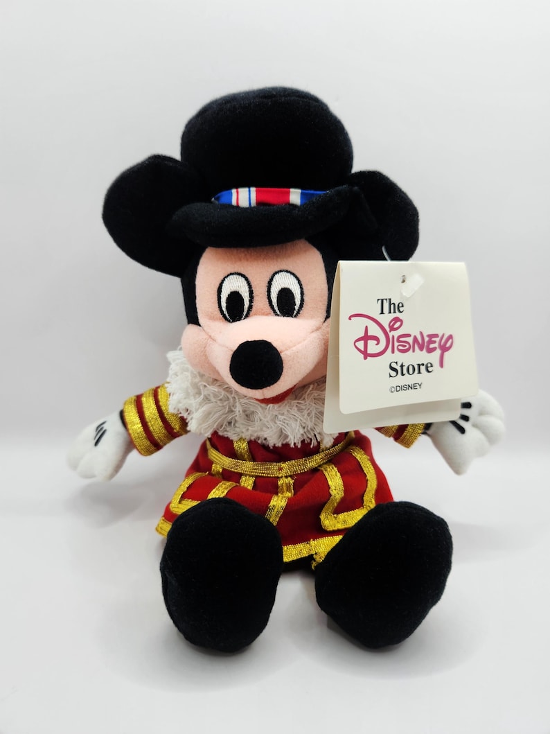 Op de afbeelding: Mickey Mouse pluche speelgoed met een rood en goud kostuum, een witte gefranjerde kraag en een zwarte hoed. Het speelgoed heeft zwarte oren en voeten, witte handschoenen en een Disney Store label.