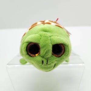 May include: A plush green turtle toy with large, glittery brown eyes and a brown and tan patterned shell. The turtle is sitting on a clear, angled display stand.