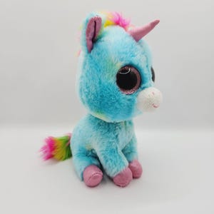 Op de afbeelding: Een blauwe en witte pluche unicorn met regenbooghaar en een roze hoorn. Het speelgoed heeft grote, glinsterende ogen en roze voeten.
