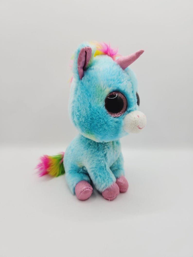 Op de afbeelding: Een blauwe en witte pluche unicorn met regenbooghaar en een roze hoorn. Het speelgoed heeft grote, glinsterende ogen en roze voeten.