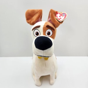 Può includere: Un peluche a forma di cane con pelo marrone e bianco, grandi occhi marroni e un naso nero. Il cane ha orecchie flosce e un collare giallo. Un'etichetta rossa a forma di cuore è attaccata all'orecchio. Il giocattolo è un personaggio del film "Pets - Vita da animali".