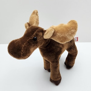 May include: A plush moose toy with dark brown fur, light tan antlers, and black plastic eyes. The stuffed animal stands on four legs and features a small white tag with red text attached to the side of its body near the back of the antlers.