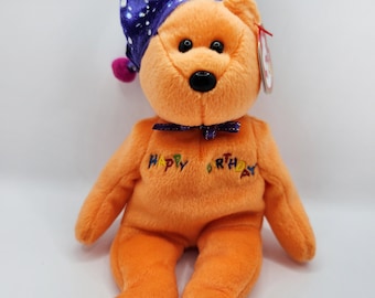 Puede incluir: Oso de peluche naranja de la marca Beanie Baby con un gorro de bufón morado y azul con pompones de colores. El oso tiene ojos negros y una nariz negra. El oso está bordado con las palabras "Happy Birthday".