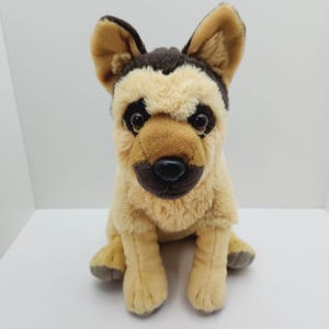 May include: A plush brown and tan German Shepherd dog toy. The dog is sitting with its paws tucked underneath it. It has brown eyes and a black nose.