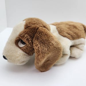 Può includere: Un peluche a forma di cane basset hound nei colori marrone, crema e bianco. Il cane di peluche ha lunghe orecchie flosce, una morbida consistenza della pelliccia e un naso nero. Il cane è sdraiato su una superficie bianca.