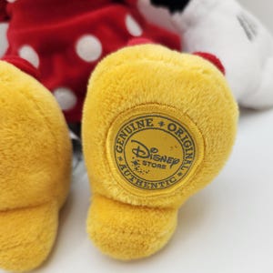 May include: A plush toy featuring a red dress with white polka dots and a white collar. The toy has yellow feet, with a circular patch on the sole that reads Genuine Original Authentic Disney Store with the Disney logo in the center.