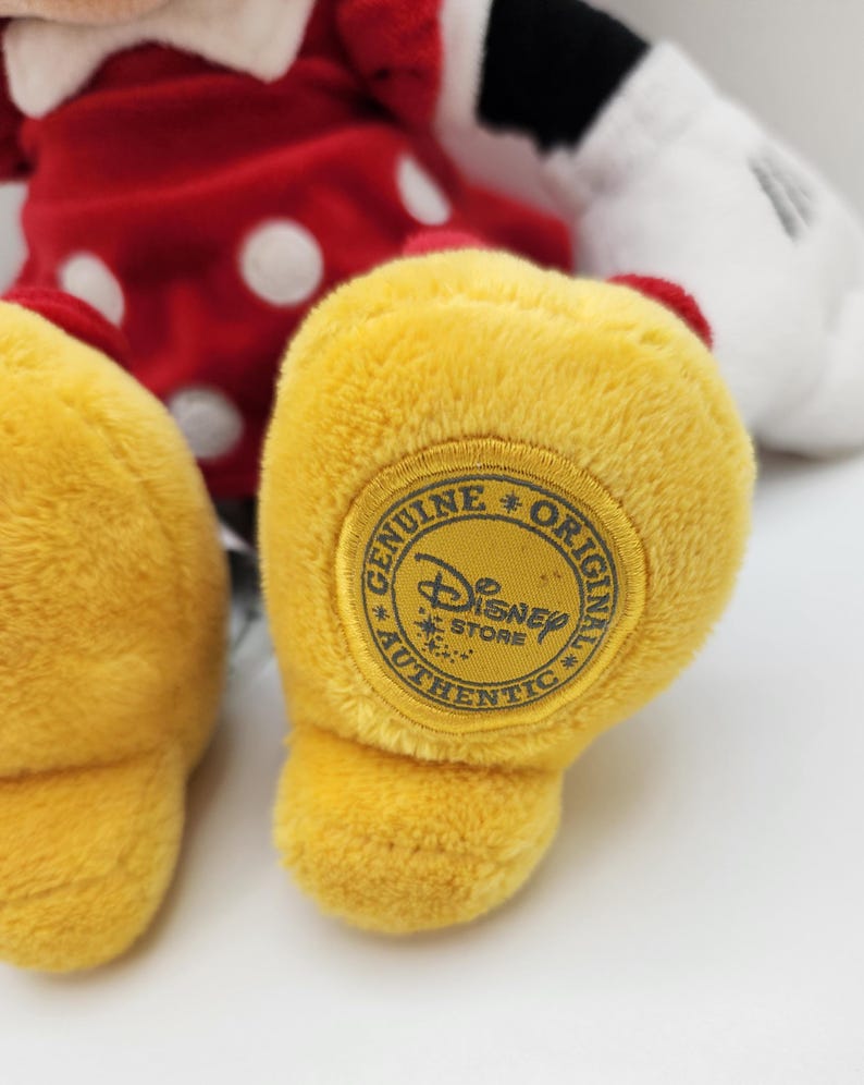 May include: A plush toy featuring a red dress with white polka dots and a white collar. The toy has yellow feet, with a circular patch on the sole that reads Genuine Original Authentic Disney Store with the Disney logo in the center.