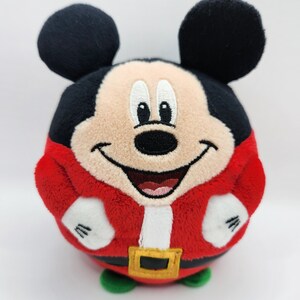 May include: A red and white plush Mickey Mouse toy wearing a Santa Claus costume. The toy is round and has a smiling face with black eyes and a black nose.