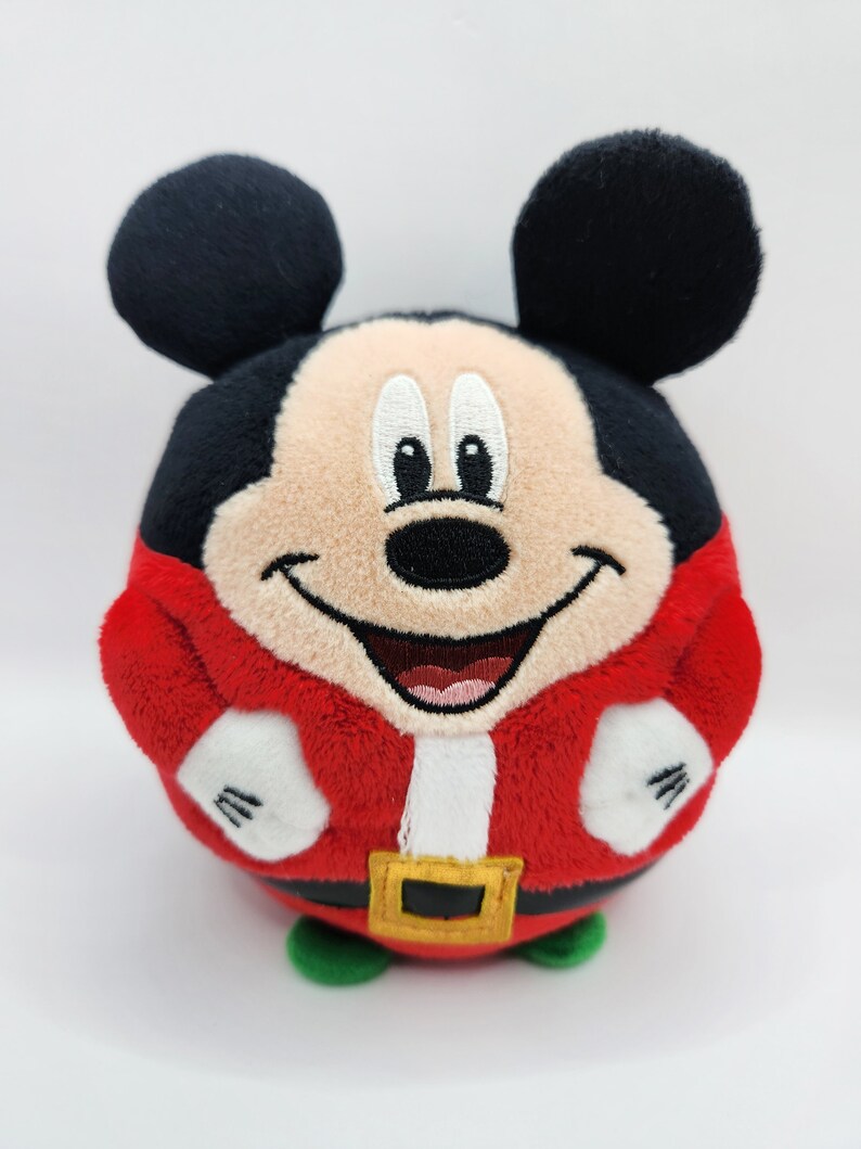 May include: A red and white plush Mickey Mouse toy wearing a Santa Claus costume. The toy is round and has a smiling face with black eyes and a black nose.