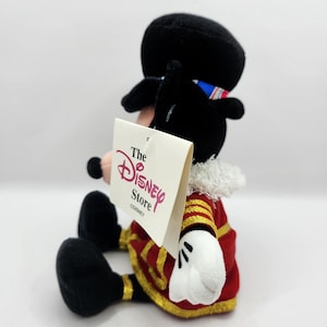 Op de afbeelding: Een pluche Mickey Mouse-figuur in een rood en goud uniform, witte handschoenen en een zwarte hoed. Het Disney Store-label is bevestigd. De figuur zit met gekruiste benen.