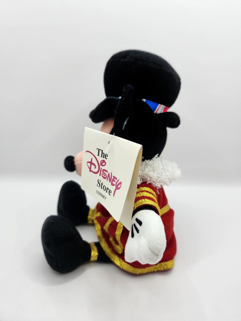 Op de afbeelding: Een pluche Mickey Mouse-figuur in een rood en goud uniform, witte handschoenen en een zwarte hoed. Het Disney Store-label is bevestigd. De figuur zit met gekruiste benen.
