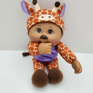 May include: A Cabbage Patch Kid doll dressed in a giraffe costume. The doll has brown eyes, a brown nose, and is wearing a purple top and brown shoes. The giraffe costume is orange and white with brown spots and purple accents.