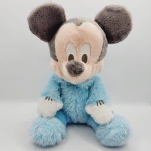 Peut inclure: Une peluche de Mickey Mouse assis. Le jouet a un corps bleu clair, des gants blancs et un visage rose avec un nez et des oreilles gris. Les yeux sont bruns. La peluche a une texture douce et duveteuse.