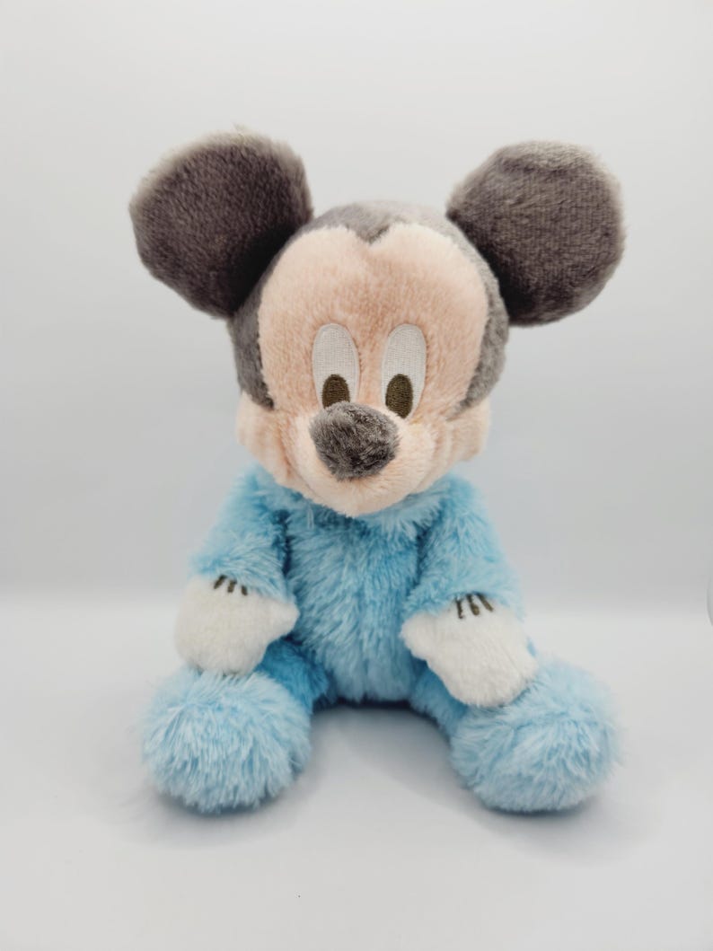 Peut inclure: Une peluche de Mickey Mouse assis. Le jouet a un corps bleu clair, des gants blancs et un visage rose avec un nez et des oreilles gris. Les yeux sont bruns. La peluche a une texture douce et duveteuse.