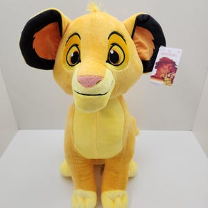 May include: A plush yellow lion cub toy with large ears, a pink nose, and big eyes. The toy has a tag that reads "Lion King". The lion is sitting upright on a white surface.