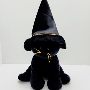 Puede incluir: Un juguete de peluche negro con forma de gato que lleva un sombrero de bruja negro con ribete dorado. El gato tiene bigotes bordados amarillos y est&aacute; sentado sobre una superficie blanca. Es un art&iacute;culo tem&aacute;tico de Halloween.