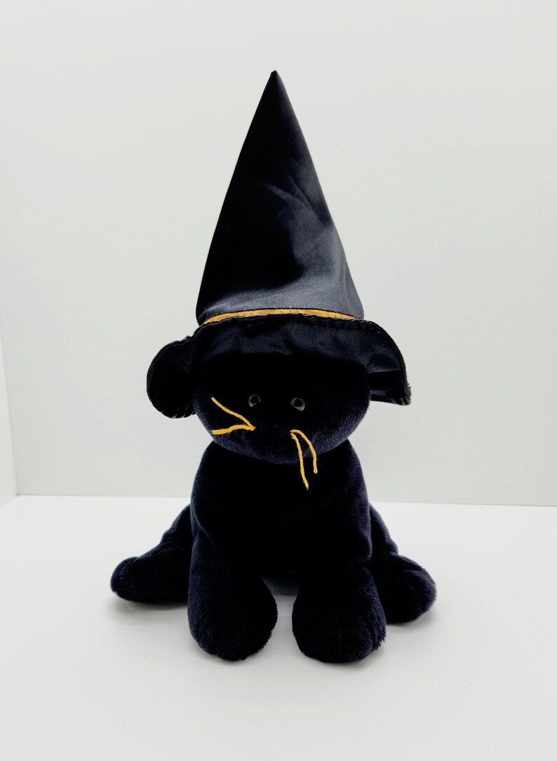 Puede incluir: Un juguete de peluche negro con forma de gato que lleva un sombrero de bruja negro con ribete dorado. El gato tiene bigotes bordados amarillos y est&aacute; sentado sobre una superficie blanca. Es un art&iacute;culo tem&aacute;tico de Halloween.