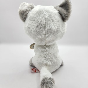 May include: A plush toy cat, viewed from the back, with soft white and gray fur. The cat has large gray ears and a fluffy tail. A small tag is attached near the tail. The toy is designed for children.