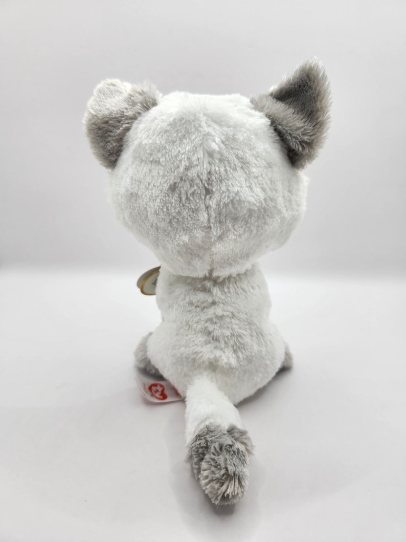 May include: A plush toy cat, viewed from the back, with soft white and gray fur. The cat has large gray ears and a fluffy tail. A small tag is attached near the tail. The toy is designed for children.