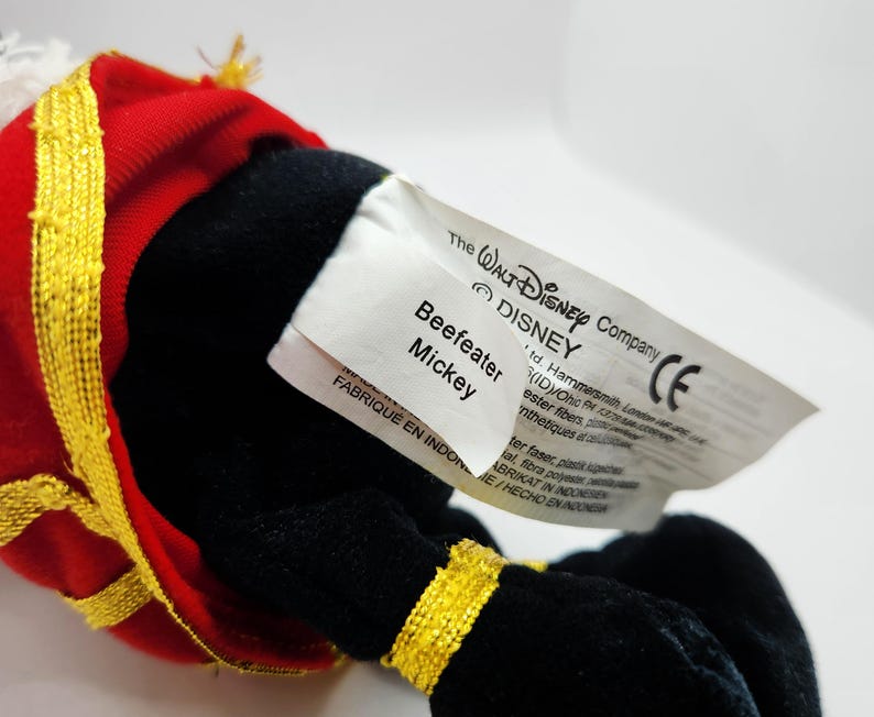 Op de afbeelding: Een Beefeater Mickey Mouse knuffel. Het speelgoed draagt een rood en goud uniform. Het label luidt "The Walt Disney Company" en "Beefeater Mickey".