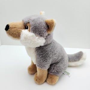 May include: A plush wolf toy featuring soft grey fur on the back and head, with tan fur on the face, chest, and paws. The toy has a black nose, brown eyes, and a white-tipped tail. A small white tag labelled Eco Nation is attached to the side.