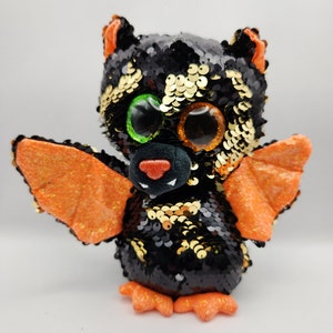 Può includere: Un giocattolo di peluche a forma di pipistrello nero e dorato con paillettes, ali arancioni e grandi occhi sbalorditivi.