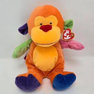 May include: A colorful plush monkey toy with orange, yellow, green, purple, pink, and blue accents. The monkey has a red heart-shaped nose and a tag attached to its arm. The toy is designed with a soft, plush material.