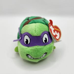 May include: A green plush toy of Donatello from Teenage Mutant Ninja Turtles. The toy has a purple mask, a brown shell, and a smiling face. A red Ty tag is attached.