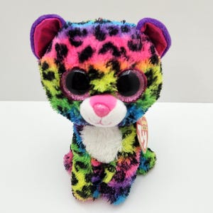 May include: A plush leopard toy featuring a vibrant rainbow-colored fur pattern with black spots. The stuffed animal has large, glittery black eyes, a bright pink nose, and purple inner ears. A small red heart-shaped tag is attached to the side.