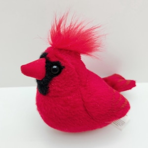 May include: A plush cardinal bird toy featuring bright red fabric, a prominent red crest of fuzzy hair on its head, a black mask around its dark eyes, and a solid red beak. The stuffed animal has a rounded body shape and a small tail.