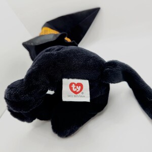 Puede incluir: Un peluche negro con forma de bruja, que lleva un sombrero negro con una banda amarilla. El juguete tiene una etiqueta blanca con un logotipo de coraz&oacute;n rojo y el texto "SHELL 100% TyLux".
