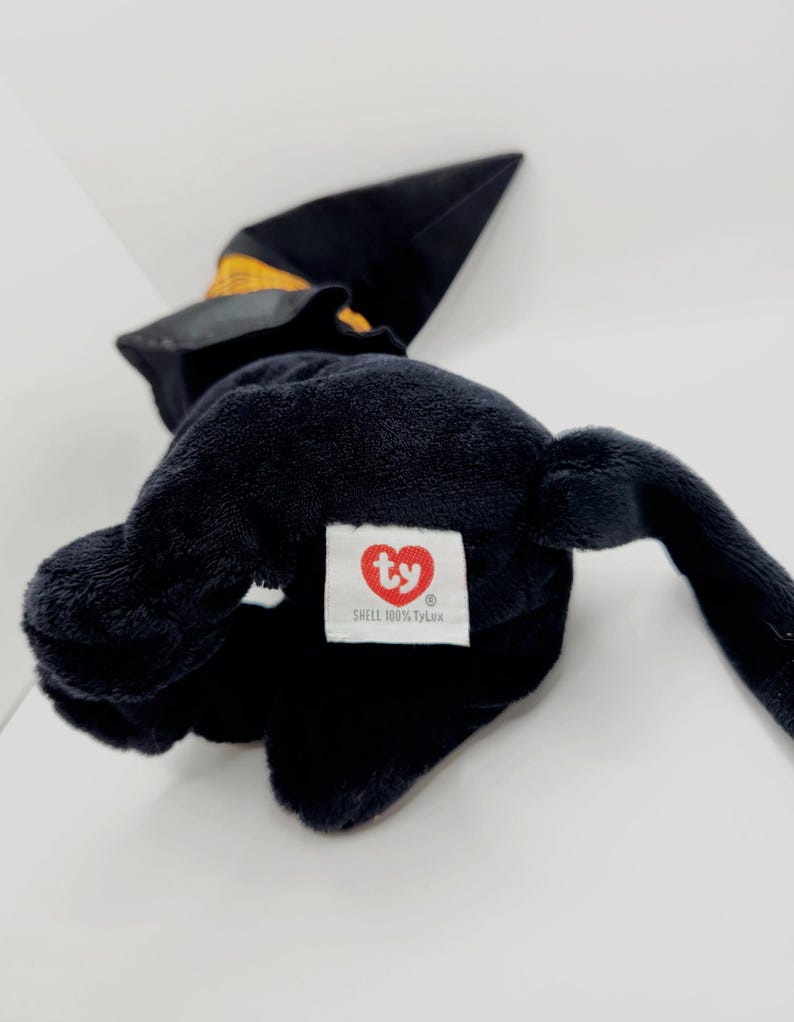 Puede incluir: Un peluche negro con forma de bruja, que lleva un sombrero negro con una banda amarilla. El juguete tiene una etiqueta blanca con un logotipo de coraz&oacute;n rojo y el texto "SHELL 100% TyLux".