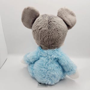 Peut inclure: Une peluche en forme de souris avec de grandes oreilles grises et un corps bleu clair. Le jouet est fait d'une mati&egrave;re douce et duveteuse et a des pattes blanches. La souris est assise sur une surface blanche.