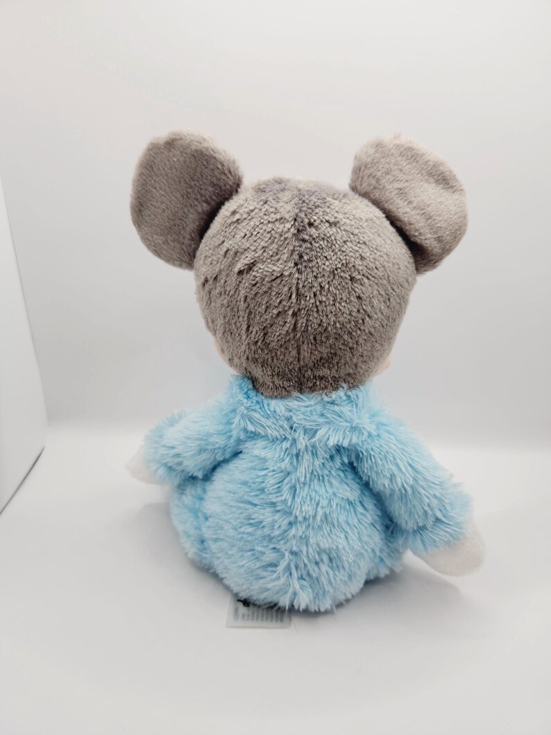 Peut inclure: Une peluche en forme de souris avec de grandes oreilles grises et un corps bleu clair. Le jouet est fait d'une mati&egrave;re douce et duveteuse et a des pattes blanches. La souris est assise sur une surface blanche.