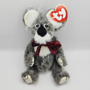 Może przedstawiać: Pluszowy miś koala w kolorze szarym z białymi akcentami i bordową kokardą. Koala ma czarny nos, okrągłe oczy i czerwoną etykietę w kształcie serca. Urocza pluszowa zabawka.