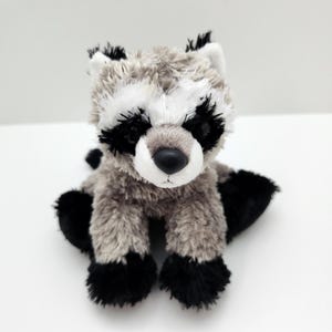 Può includere: Un giocattolo di peluche a forma di procione con pelliccia grigia, bianca e nera. L'animale di peluche ha una maschera nera intorno agli occhi, zampe nere e una coda nera. Il giocattolo è su una superficie bianca.