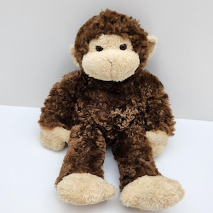 Peut inclure: Une peluche singe avec une fourrure texturée marron foncé sur le corps et la tête, contrastée par une fourrure beige clair sur le visage, les oreilles, les mains et les pieds. L'animal en peluche a deux petits yeux ronds et un museau cousu.