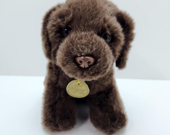 May include: A plush, brown Labrador puppy dog with a soft, textured fur. The puppy has a small, round nose, dark eyes, and floppy ears. A gold-colored tag hangs from its neck. The stuffed animal is sitting upright on a white surface.
