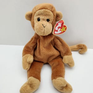 Peut inclure: Un singe en peluche marron avec un visage, des mains et des pieds d'une teinte beige plus claire. Ce jouet en peluche possède des yeux en plastique noir et une longue queue. Une étiquette rouge en forme de cœur avec les lettres ty blanches est attachée à l'oreille gauche du singe.